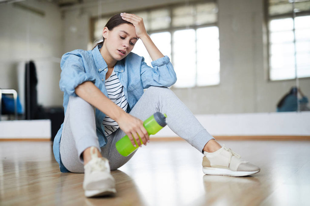 Exhausted active woman with bottle of water sitting on the floor of modern dance studio and having break - Photo, Image