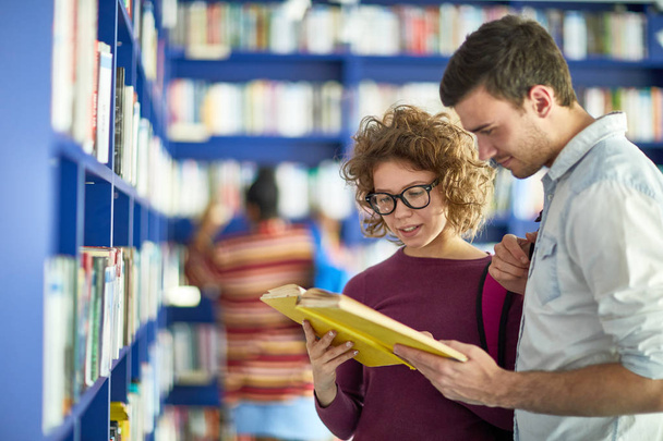 positive junge Studenten in lässiger Kleidung stehen am blauen Regal und lesen gemeinsam Bücher, während sie in der Bibliothek die notwendigen Informationen finden - Foto, Bild