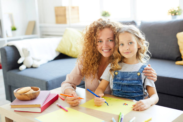 junge fröhliche Frau und ihre Tochter sitzen am Tisch und zeichnen mit Textmarker im Wohnzimmer - Foto, Bild