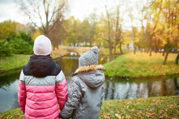 Back view of two school friends in warm jackets and beanies holding by hands while standing in front of waterside on autumn day - Valokuva, kuva