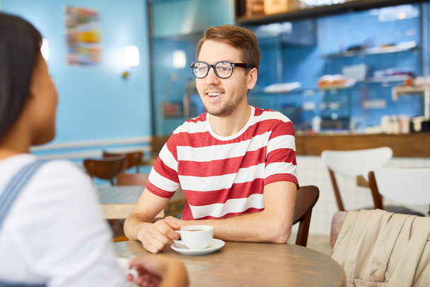 Joyeux jeune homme avec une tasse de café assis à table devant un collègue lors de leur conversation au café
 - Photo, image