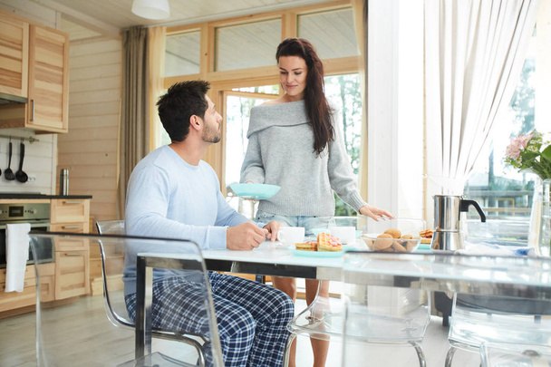 Content careful attractive young wife with black hair carrying breakfast to husband and putting plate on table, handsome husband waiting for his breakfast in kitchen - Photo, Image