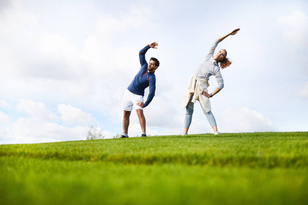 Active guy and girl in sportswear doing side bend exercise while standing on green field - Photo, Image