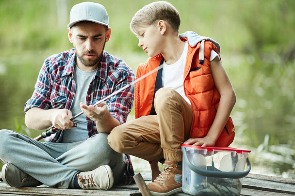 Concentrado pai mostrando seu filho como amarrar um verme em uma vara de pesca enquanto eles pescam
 - Foto, Imagem