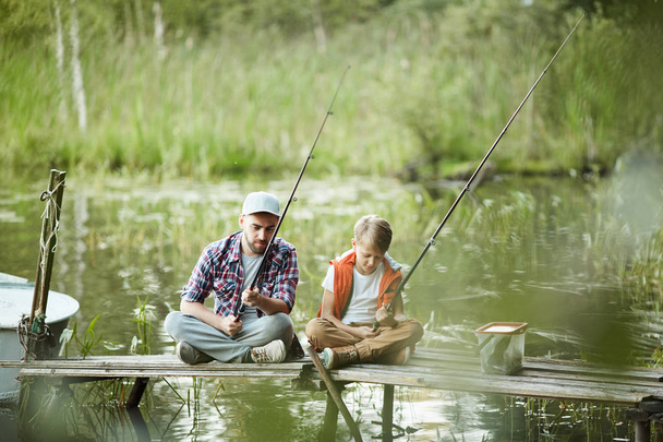 Papá e hijo sentados en el muelle con cañas de pescar en el río en el día de verano
 - Foto, imagen