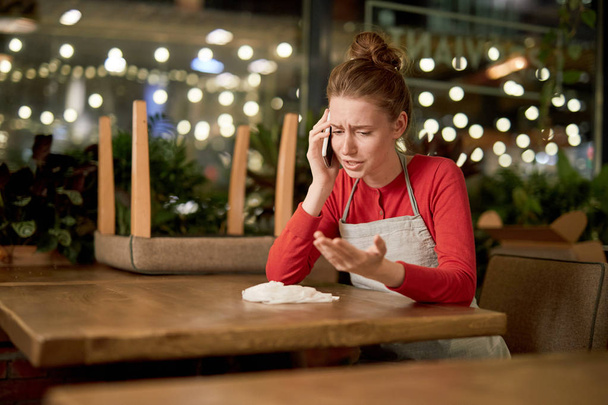 Upset waitress sitting by table and talking to someone by smartphone at the end of working day - Foto, Bild