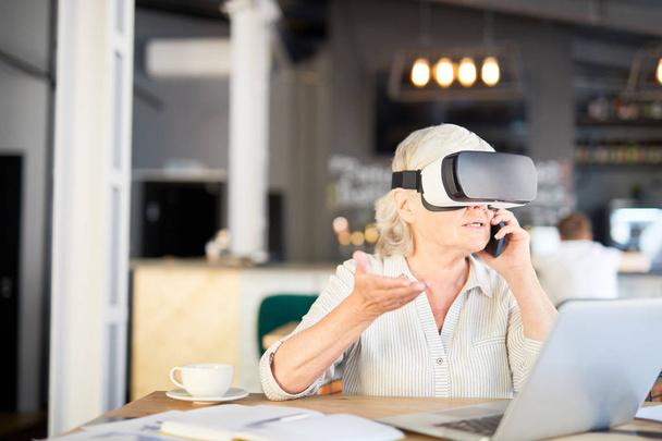 Зрелая деловая женщина сидит в кафе перед ноутбуком, разговаривает по смартфону и смотрит виртуальную презентацию в наушниках vr
 - Фото, изображение