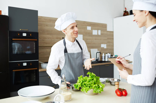 Zufriedener junger Mann in Koch-Uniform besteht praktische Prüfung beim Kochkurs: Er erzählt Prüfern von Salatrezept - Foto, Bild