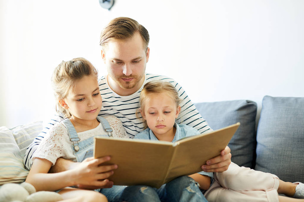 Ο νεαρός πατέρας και οι δύο κόρες του διαβάζουν βιβλία με ιστορίες ενώ μένουν στο σπίτι. - Φωτογραφία, εικόνα