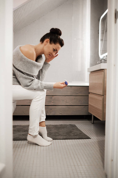Upset shocked attractive young woman with hair bun sitting on toilet bowl in bathroom and looking at pregnancy test, she not believing her eyes - Фото, изображение