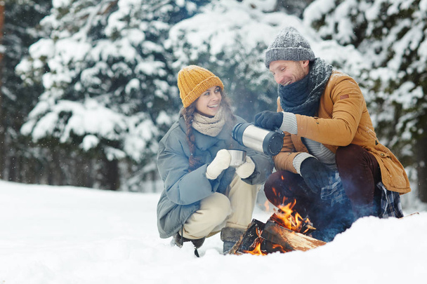 Happy excited young couple in warm jackets drinking hot tea and sitting by fire while enjoying camping in winter forest - Photo, Image