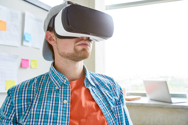 Poważny uczeń z brodą noszenie gogli VR oglądania wideo edukacyjnych i siedzi w nowoczesnym biurze - Zdjęcie, obraz