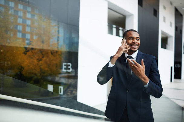 Feliz joven empresario africano en traje hablando por teléfono inteligente en el entorno urbano
 - Foto, Imagen