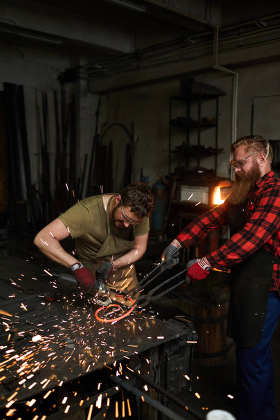 Two blacksmiths processing spiral molten metal workpiece with electric handtool on anvil - Fotoğraf, Görsel