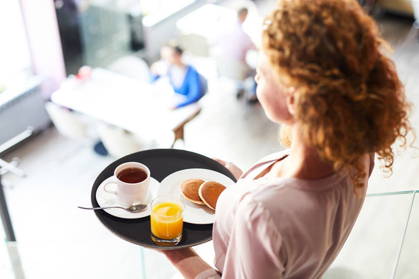 Mujer joven llevando panqueques, té y jugo en bandeja mientras se mueve a una de las mesas en la cafetería
 - Foto, imagen