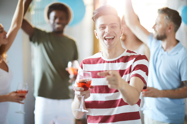 Lachende kerel met drankje naar je wijst terwijl je plezier hebt op feest met dansende vrienden op de achtergrond - Foto, afbeelding