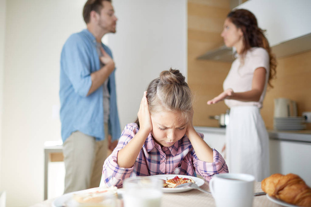 Gestrest meisje bedekt haar oren terwijl leunend over geserveerd tafel bij het ontbijt op de achtergrond van haar ouders ruziën - Foto, afbeelding