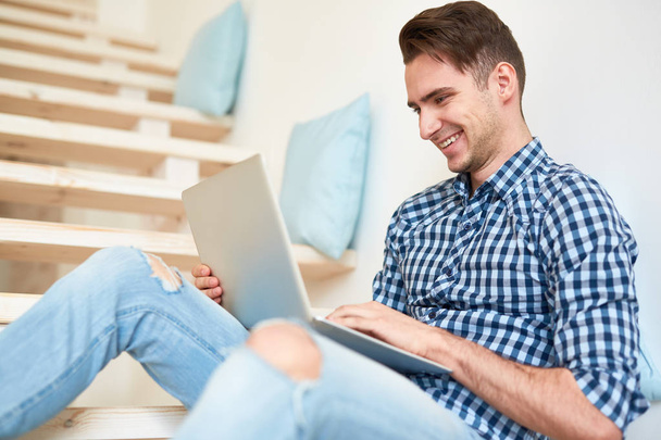 Jonge lachende student of ontwerper zittend op de trap met laptop voor en zoekend in het net - Foto, afbeelding