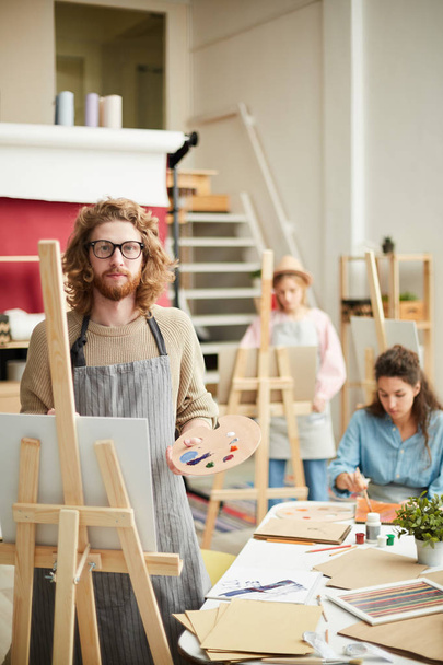Serious young man with palette standing by easel and looking at camera in working environment - Photo, Image