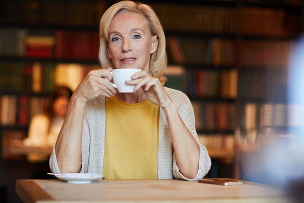 Happy blonde aged woman looking at you while enjoying coffee by tablein cozy cafe - Photo, Image