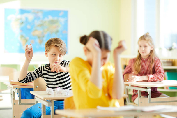 Θυμωμένος σχολείο τραμπούκος ρίχνοντας χαρτί μπάλα στο νέο κορίτσι και κάθονται στο γραφείο στην τάξη γεωγραφία, εκφοβισμός στο σχολείο έννοια - Φωτογραφία, εικόνα