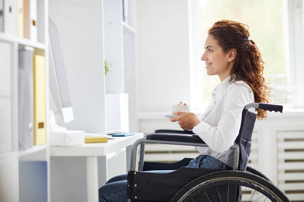 Ofisteki genç bayan tekerlekli sandalyede bilgisayarın önünde oturuyor, çay içiyor ve internet yayını izliyor. - Fotoğraf, Görsel