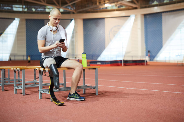 Mistrz Paraolimpijski młodych lub konkurenta ze słuchawkami, siedząc na ławce podczas przewijania w smartfonie do listy odtwarzania przy zerwaniu - Zdjęcie, obraz
