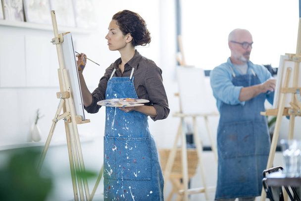 Jonge vrouw in denim vuil schort schilderen met penseel en kleuren in studio van kunst - Foto, afbeelding