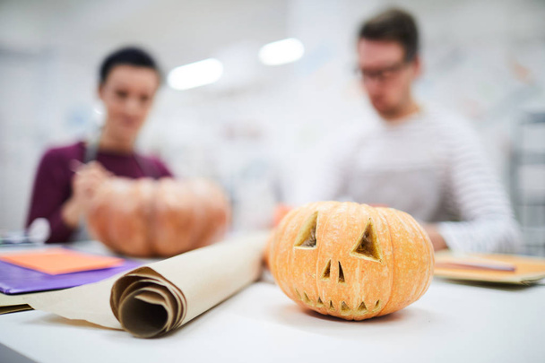 Nahaufnahme von geschnitztem Halloween-Kürbis und Pergamentpapierrolle auf dem Tisch, Bastler arbeiten im Hintergrund - Foto, Bild
