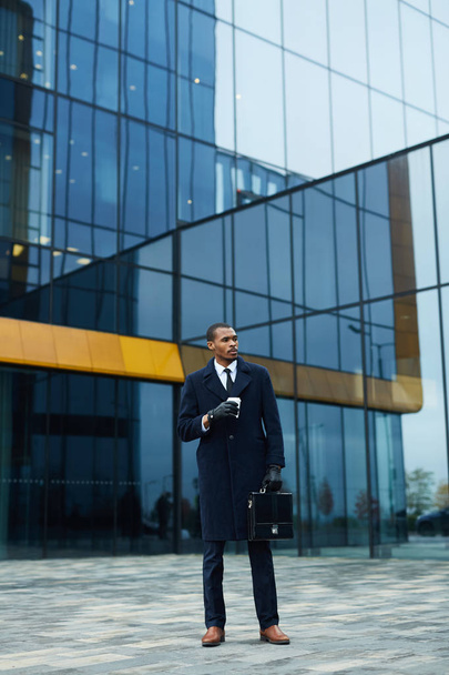 Jeune homme d'affaires afro-américain élégant avec mallette et boisson debout près du centre de bureau moderne
 - Photo, image