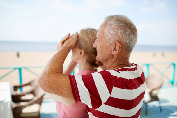 Vanhempi mies peittää vaimonsa silmät ennen hänen kesälomakohdettaan ja rantaa
 - Valokuva, kuva