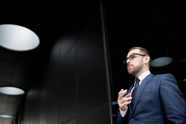 Νεαρός επιτυχημένος άνδρας με επιχειρηματικό φόρεμα, γυαλιά καπνίσματος και κοιτάζοντας μακριά  - Φωτογραφία, εικόνα