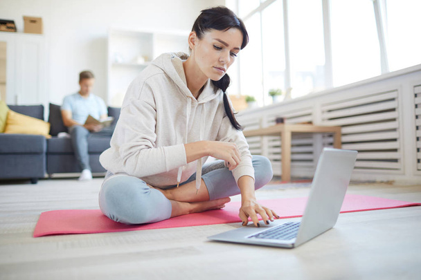 Mujer activa en busca de entrenamiento en línea mientras está sentada en el suelo frente a la computadora portátil en la sala de estar
 - Foto, imagen