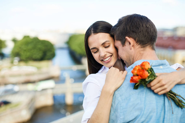 Happy tender young couple hugging each other while dancing on city roof and enjoying romantic date, pretty lady holding small bouquet - Фото, изображение