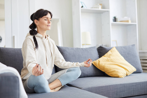 Mujer joven sentada en pose de loto en el sofá con los ojos cerrados durante el entrenamiento de yoga
 - Foto, imagen