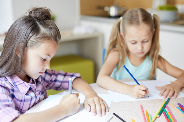 Kaksi söpöä tyttöä istuu pöydän ääressä ja piirtää kuvia värikynillä oleskellessaan kotona
 - Valokuva, kuva