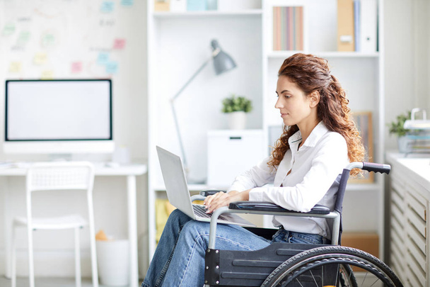 Молодая женщина в повседневной одежде сидит в инвалидной коляске с ноутбуком впереди и ищет информацию в сети
 - Фото, изображение