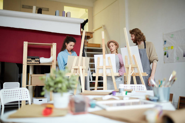 Group of young artists standing in front of their easels, painting and consulting in working process - Photo, Image