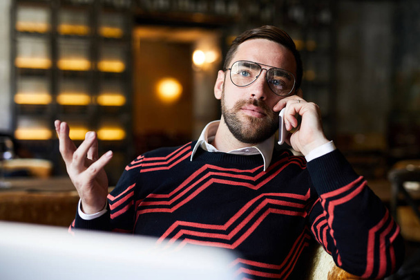 Jovem homem de negócios caucasiano em casualwear falando por smartphone enquanto sentado no café
 - Foto, Imagem