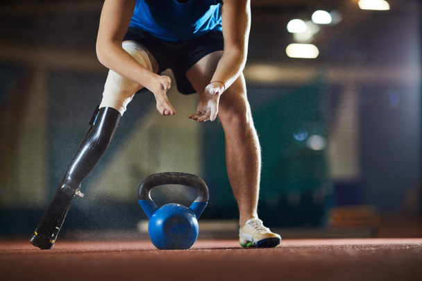 Young paralympic athlete leaning over heavy kettlebell before taking and lifting it during workout - Photo, Image