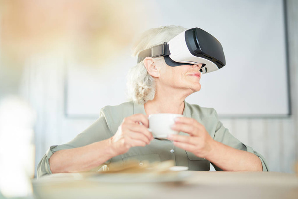 Donna anziana moderna con auricolare realtà virtuale seduta a tavola e che beve tè o caffè
 - Foto, immagini