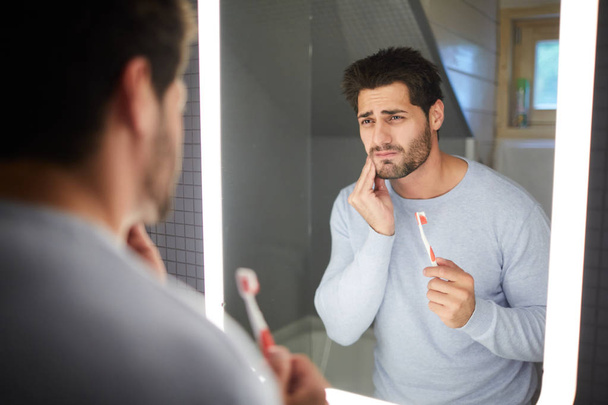 Sad frowning handsome brunette young man with beard touching cheek and looking into mirror in bathroom while getting bad tooth during cleaning teeth - Фото, зображення