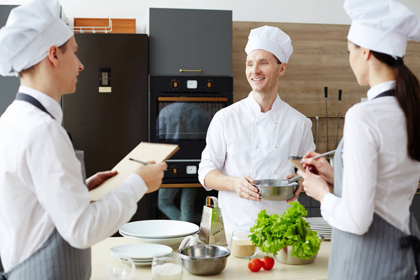 Cheerful chef bonito otimista em tigela chapéu segurando com batedor ao compartilhar ideias culinárias enquanto prepara nova receita, juntamente com os colegas na cozinha comercial
 - Foto, Imagem