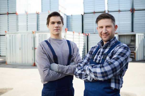 Two young delivery men with arms crossed on chest looking at camera on background of containers - Фото, изображение