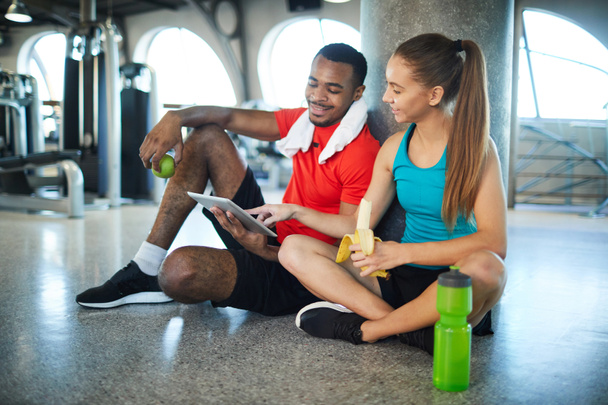 Молодой активный мужчина и женщина перекусывают во время перерыва между тренировками и просмотром онлайн видео в планшете
 - Фото, изображение