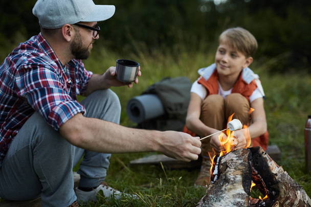 キャンプ旅行中に火の近くで息子と話すお茶とマシュマロを持つ父 - 写真・画像