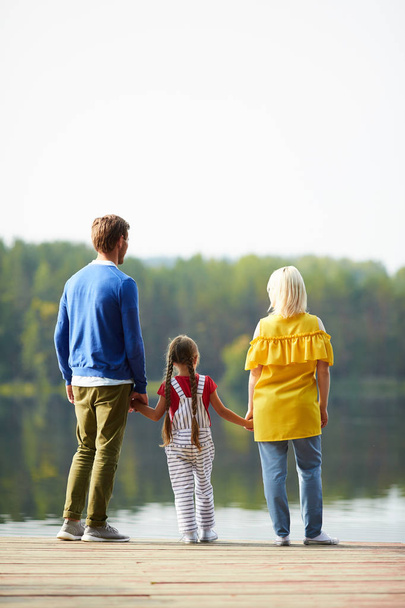 Rear view of family of three standing on pontoon and looking at river or lake while enjoying rest in natural environment - Photo, Image