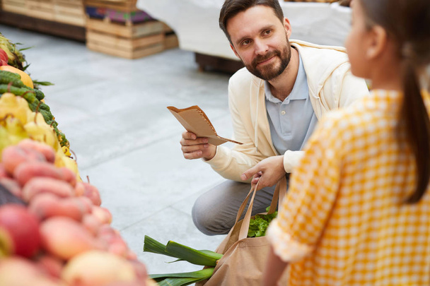 Content handsome bearded father crouching and holding full bag and shopping list while listening to daughter while they buying organic food - 写真・画像