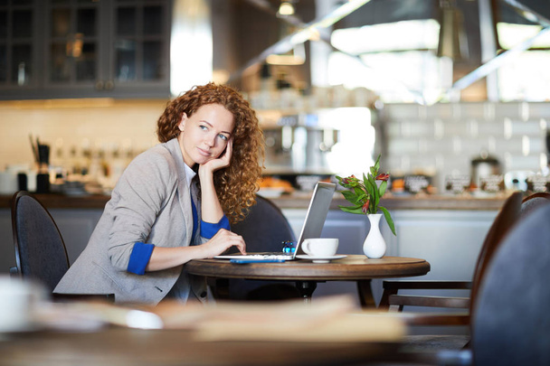 Nachdenkliche junge Frau mit welligem Haar sitzt am Tisch vor dem Laptop und entspannt im Café - Foto, Bild