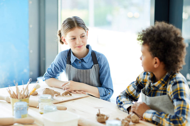Mladá dívka mluví se svým spolužákem o kreativních nápadů, zatímco sedí u stolu na lekci ve studiu řemesel - Fotografie, Obrázek